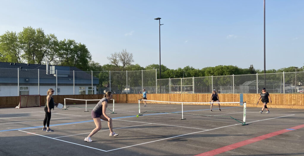 Four pickleball players face-off in the Riverview Community Centre's new paved rinks, using removable nets, and painted on lines. 